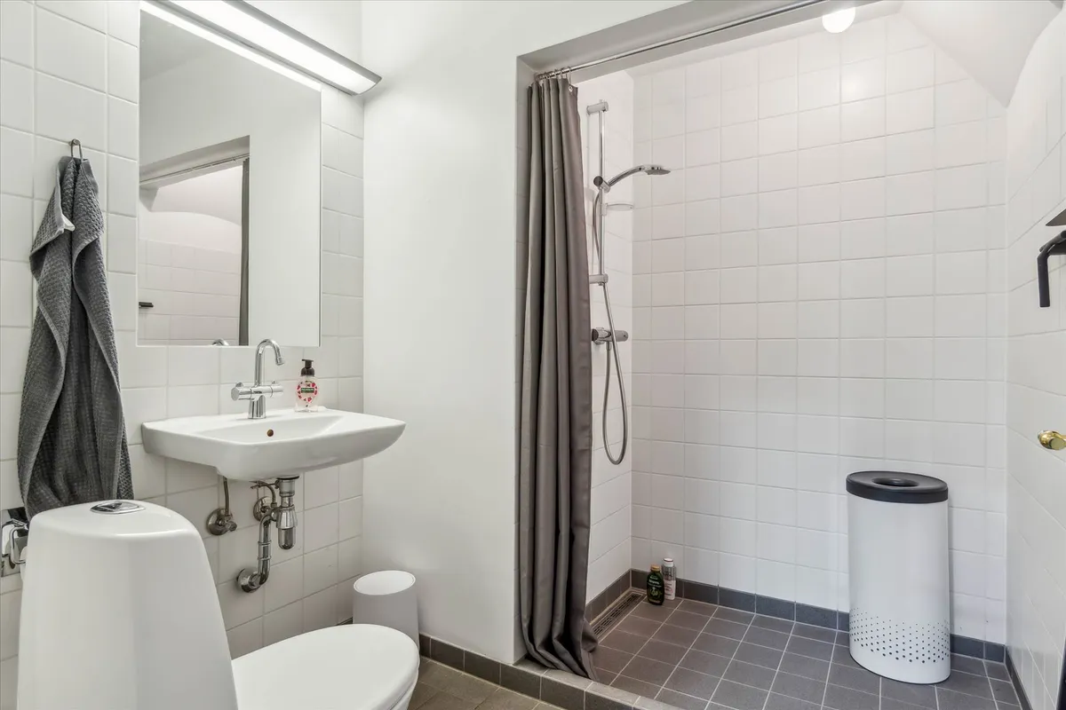 Et moderne hvidt badeværelse med brusebad, hvid toilet, hvidt spejl, hvidt håndvask, og en hvid papirkurv med sort låg