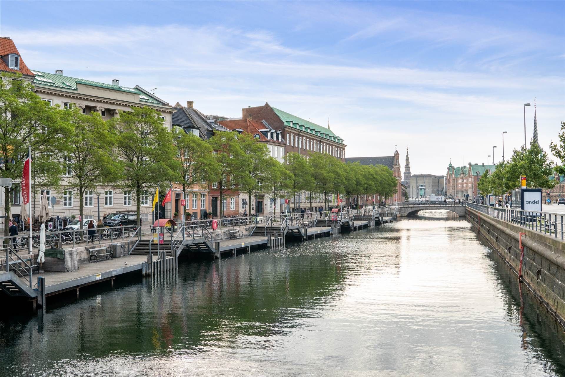 Kanal med grønne træer, huse og bro. Mennesker går på gågaden, og biler er parkeret. Solskin og blå himmel.