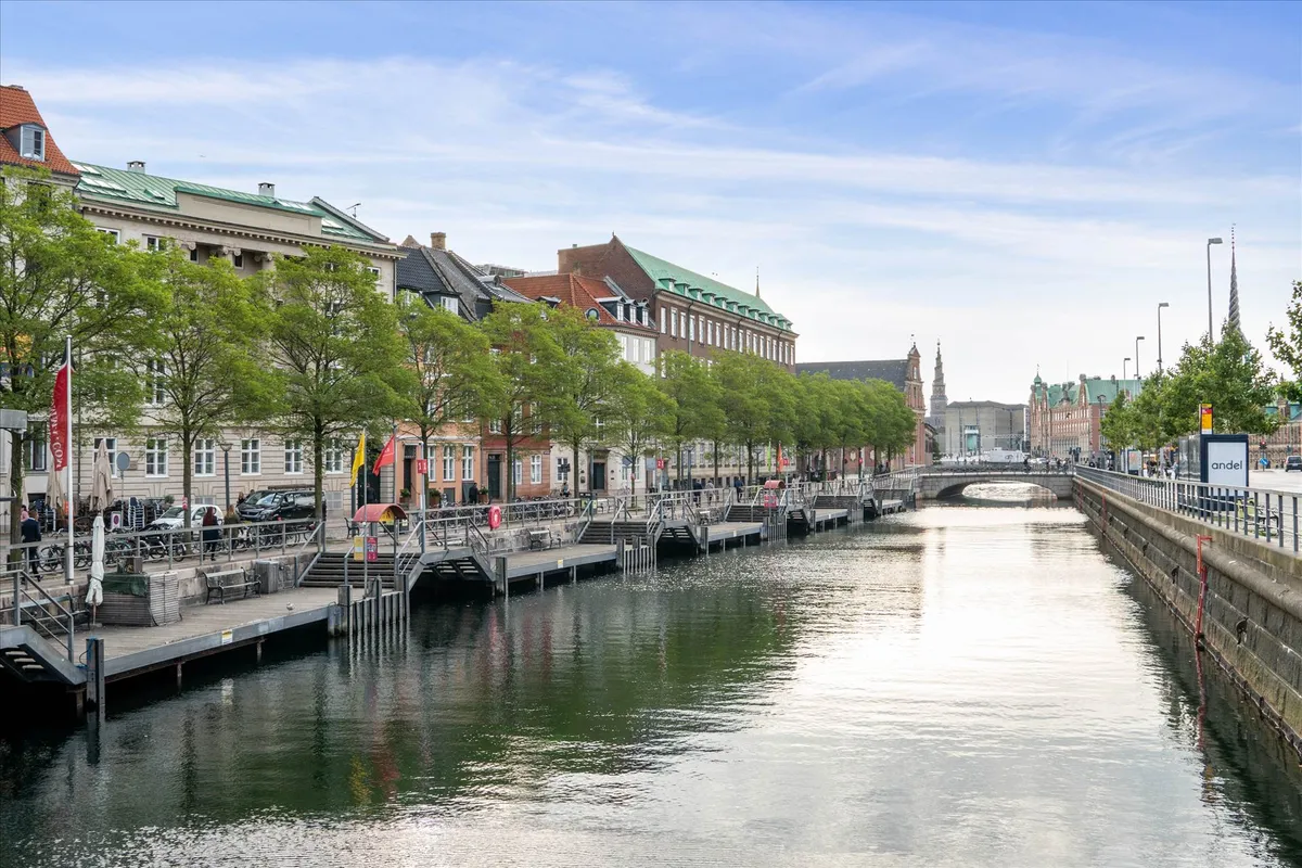 Kanal med grønne træer, huse og bro. Mennesker går på gågaden, og biler er parkeret. Solskin og blå himmel.