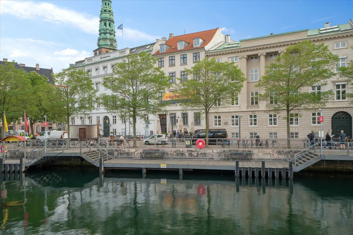 En bred kanal med flotte grønne træer og moderne bygninger i baggrunden, bl.a. Christiansborg Slot.