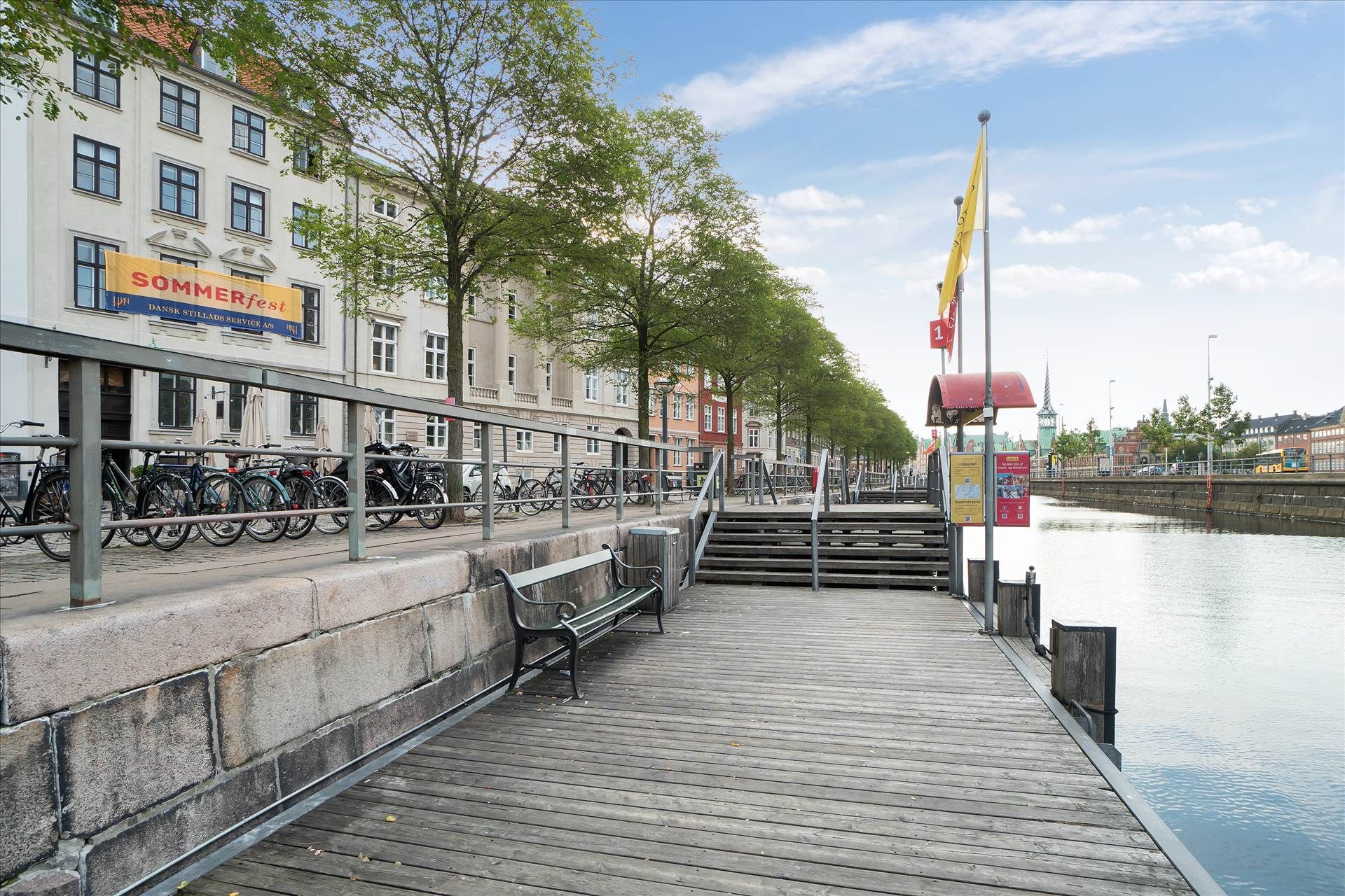 Langs Slotsholmskanalen står en bænk på en træbro med trapper og skraldespand, og i baggrunden er et bygningskompleks med et
