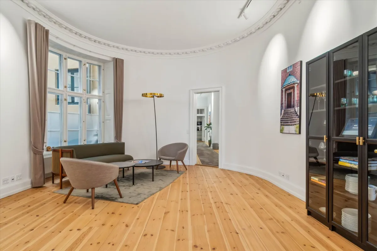 Moderne, lyse kontorlokaler med højloft, hjørnesofa, stol, kaffebord, gulvtæppe og lysegule vægge med kunstværk.