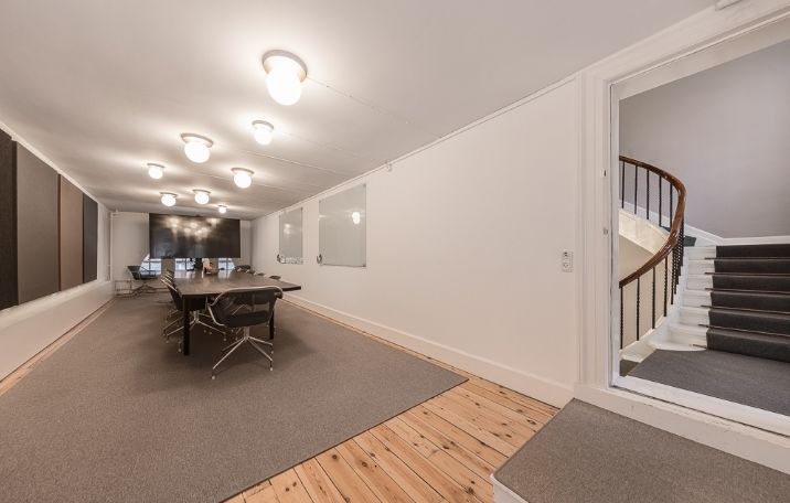 Moderne mødelokale med lyse loftslamper, trægulv og gulvbelægning langs væggen og trappen