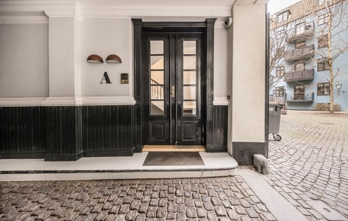 Stort sort glasdøre på en høj hvid mur, to messinglys og et skraldespand i baggrunden