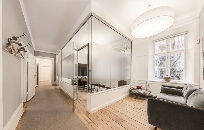 Kontor med højloftede lokaler og høje paneler. Væggene er malet hvidt. Der er en stor glasvæg og trægulv. Der er en sofa og