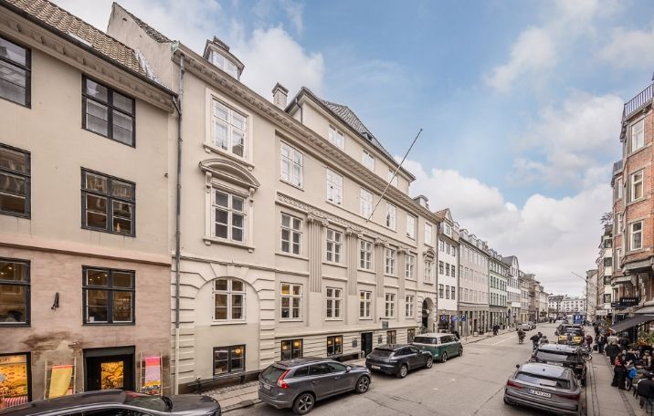 Flot bygning med store vinduer og røde tagtægler placeret ved en travl gade med parkerede biler og gående mennesker