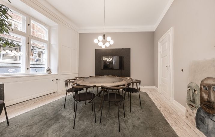 Rundt konferencebord med stole og en stor tv-skærm i et højloftet kontor med lyskrone og lysevinduer