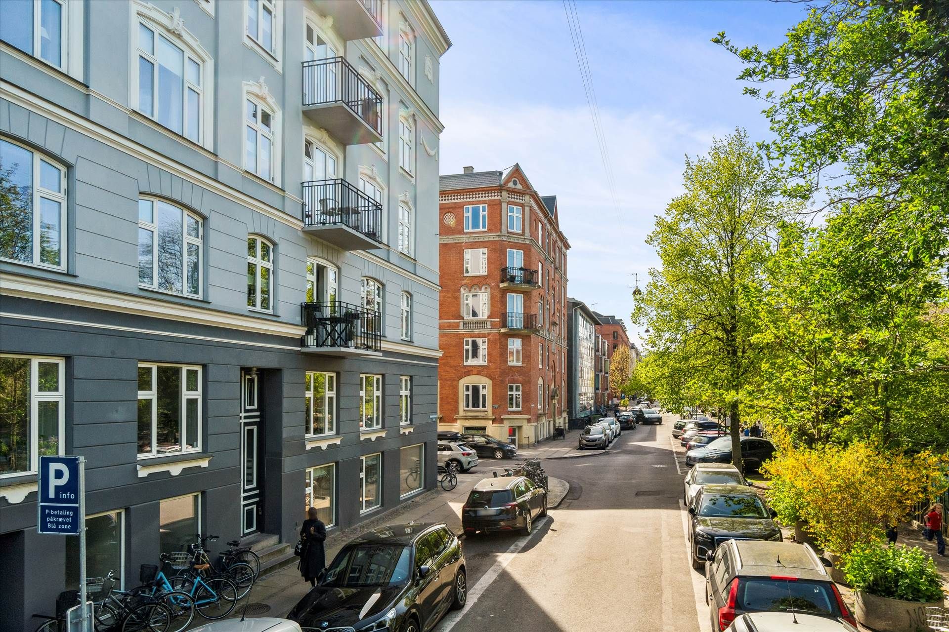 Parkerede biler langs en gade med gamle bygninger, træer og buske samt en person, der går på fortovet.