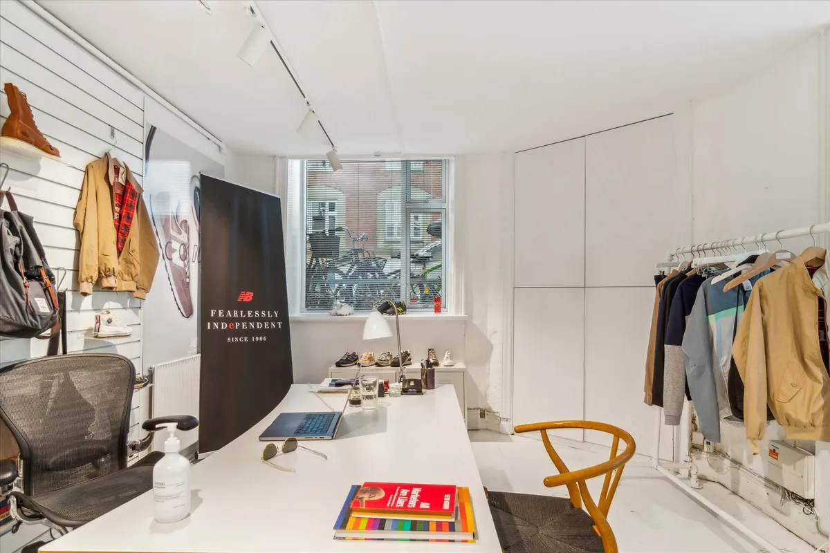 Kontor med hvidt gulv og væg, høj hylde med New Balance varer, sort stol og skrivebord med laptop, sæde og lampe