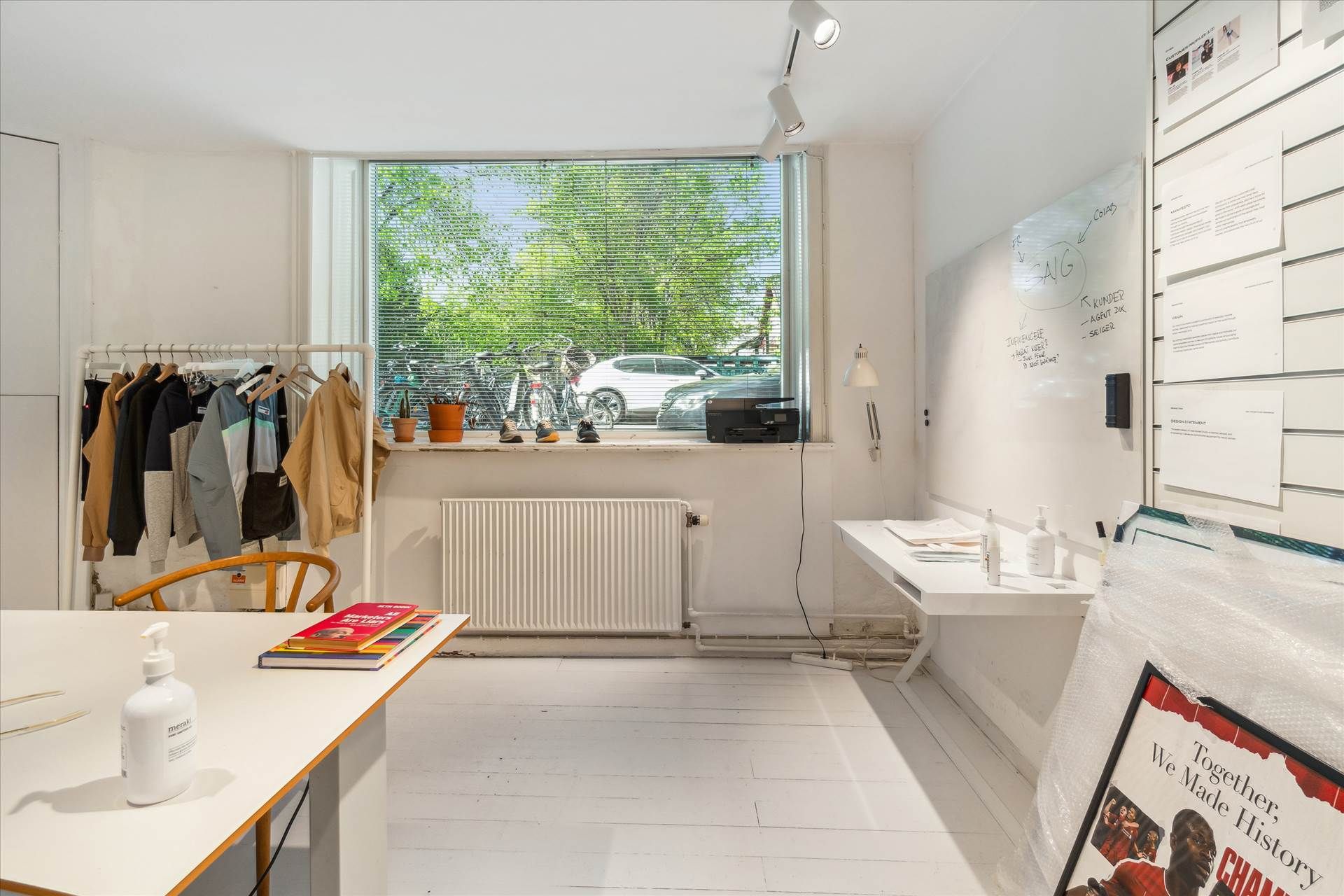 Kontor med hvide vægge, højloft, hængende tøj, radiator, bøger, mærskvande og plakat i ramme samt plante og cykler udenfor