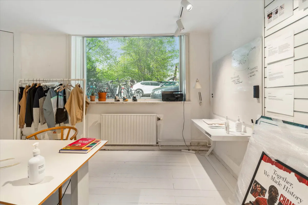 Kontor med hvide vægge, højloft, hængende tøj, radiator, bøger, mærskvande og plakat i ramme samt plante og cykler udenfor