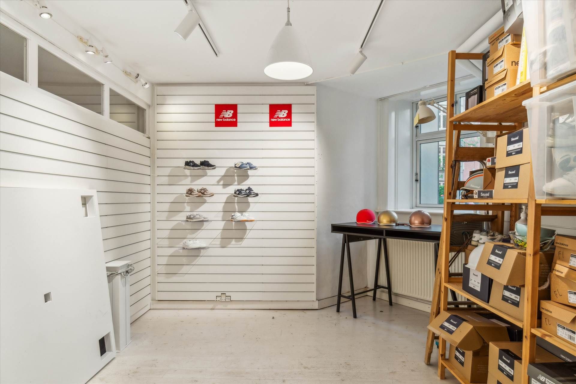 En butik med New Balance sko, støvler og hjelme. Hældende bord med briller og flasker. Skabe med sko i kartoner. Vindue med