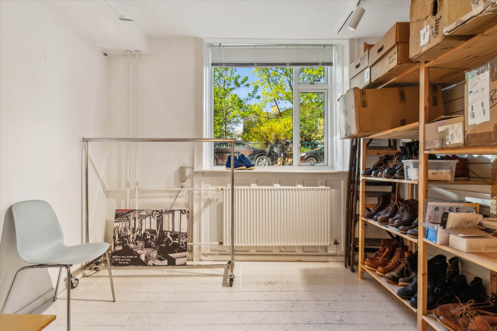 En række sko står på hylder og i kasser, der er opstillet på en hvid gulv. En klapvogn med hjul og en hvid stol står i