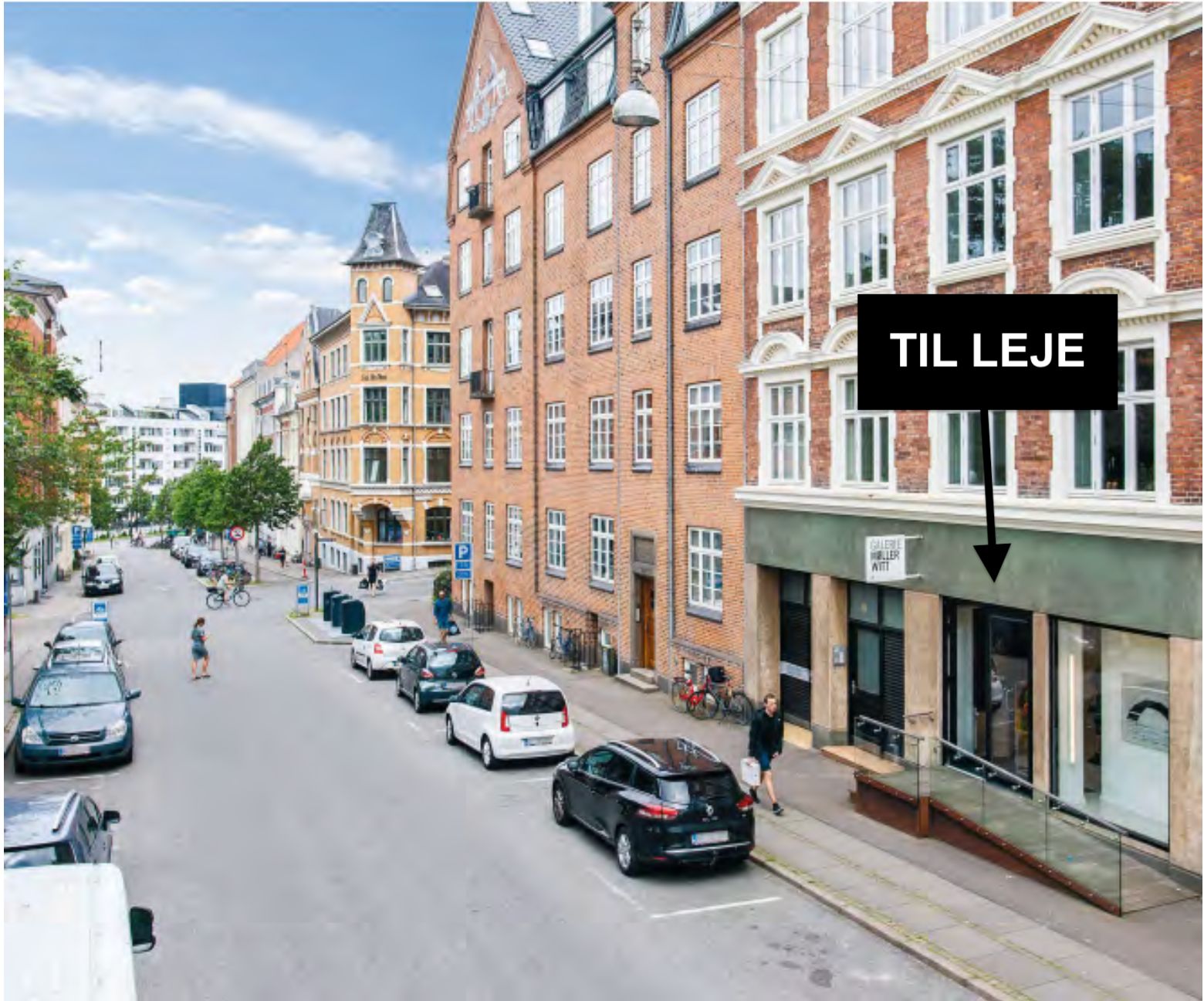 Til leje skrift på bygning og en person i gangen med en kasse, og biler parkeret langs en gade med folk og cykler
