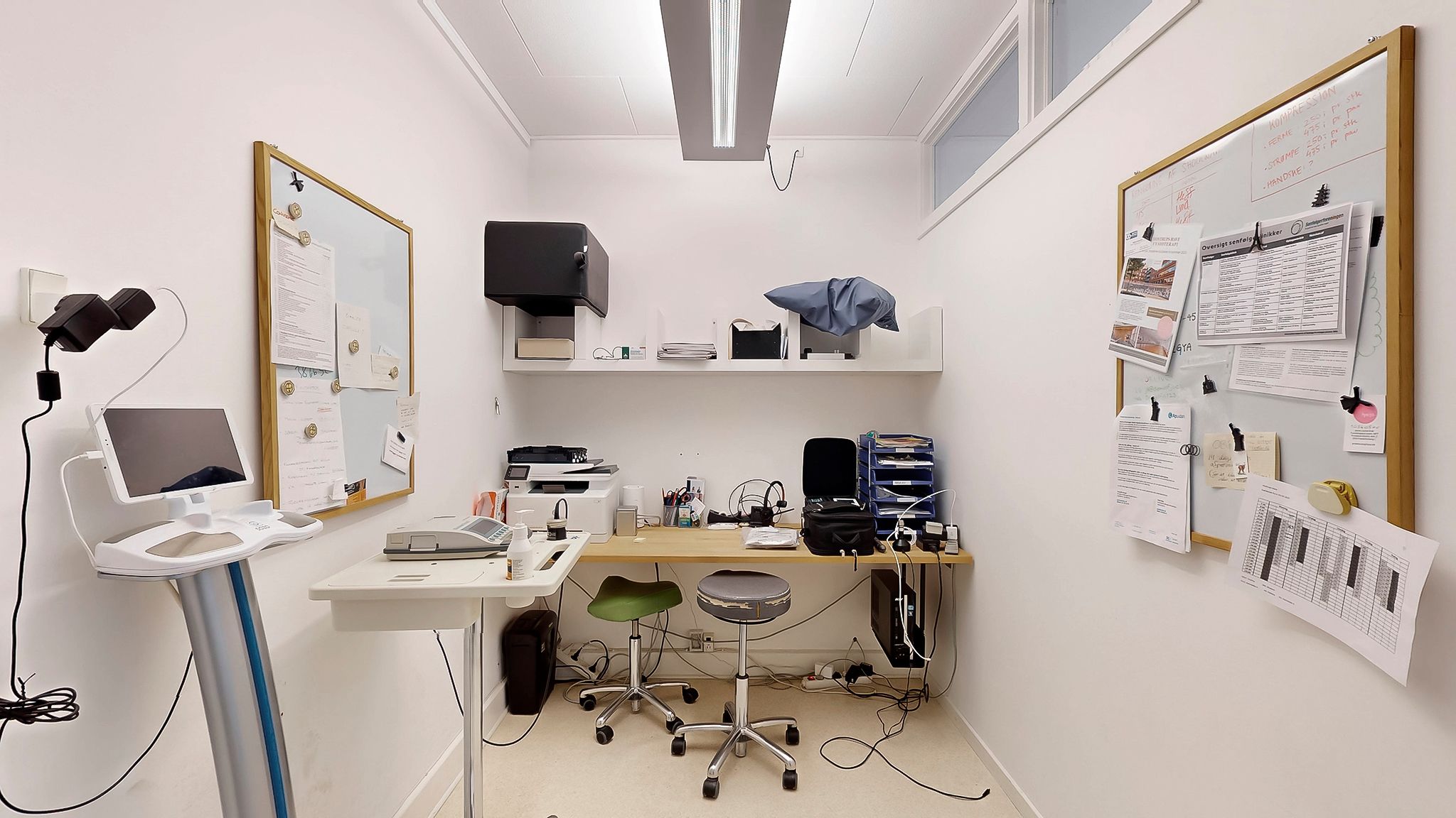 Et kontorlokum med en printer, skrivebord og computer. To stole og et bord med en trækasse på. Væg med vippeskilte og