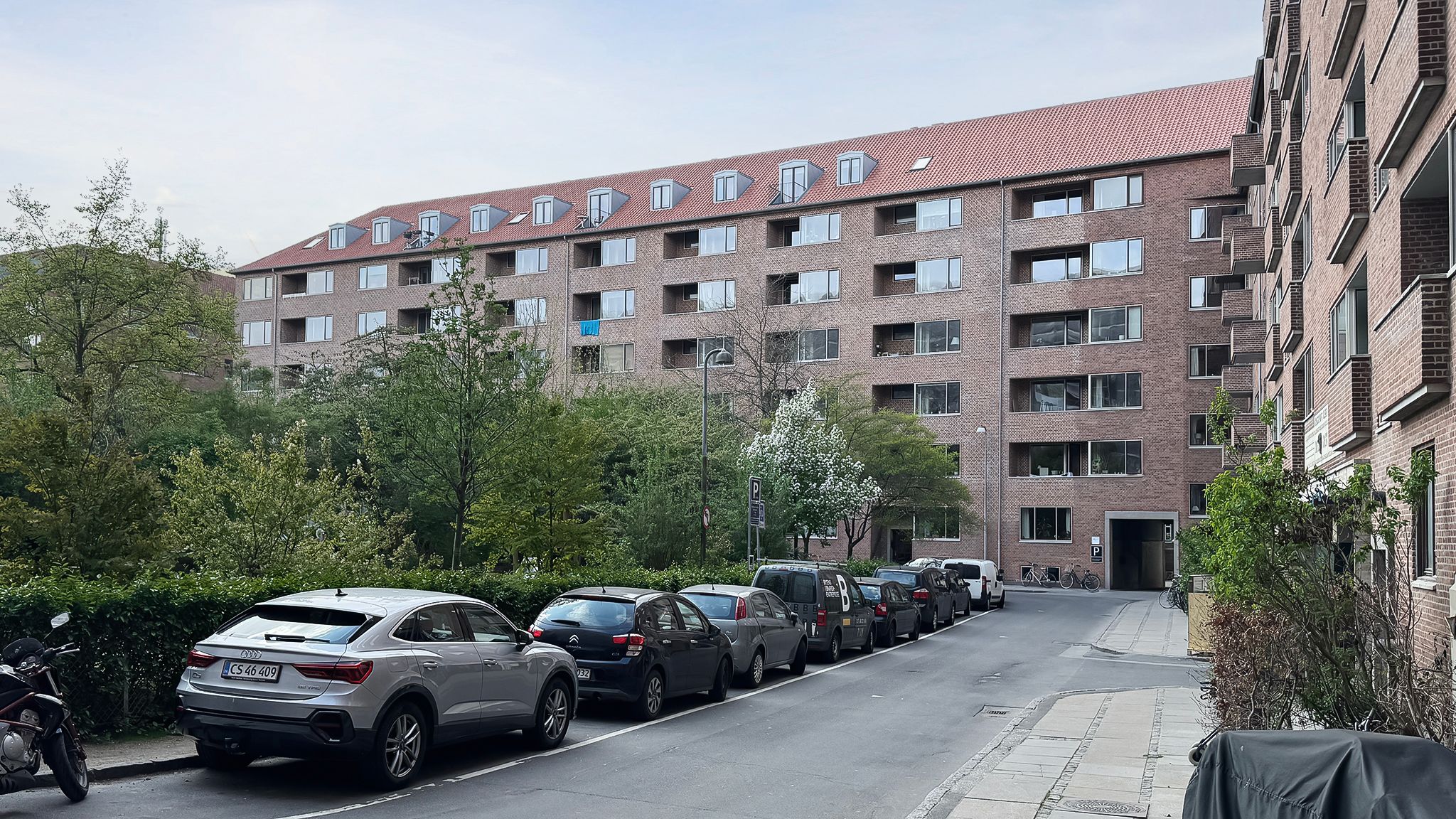 Parkerede biler langs en gågade med gamle træer, en stor lejlighedsbygning med røde tag, og en skovet grøn plante med