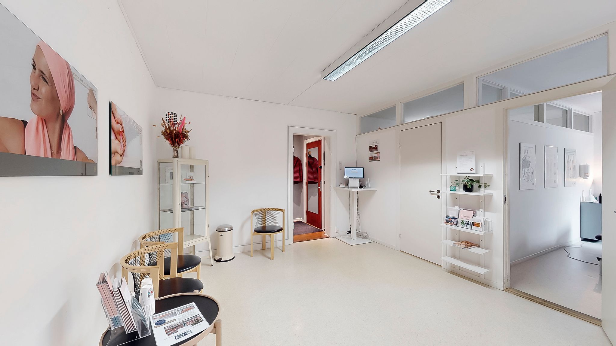 Inde i det hvide lokale er der stolpe med bøger og en plante, samt en computer på et bord og et sæt stole og bord