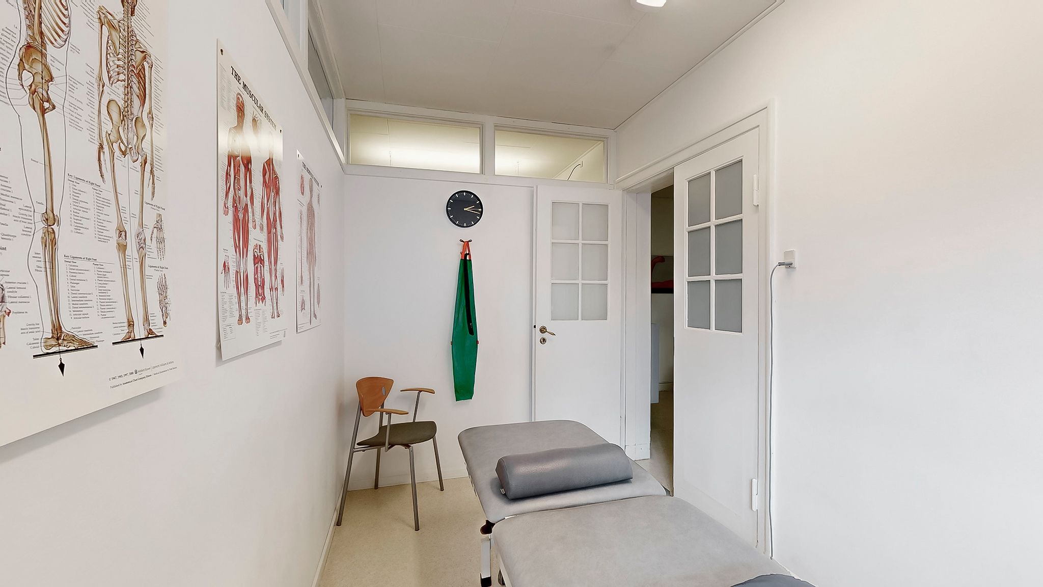 Et lille lægeværelse med en hvid væg, to lægestolar, en sofa, et ure og to skilte med menneskelige kroppens muskler