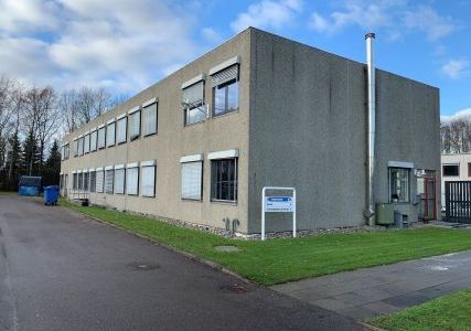 Moderne kontorbygning med vinduer og døre. Bygningen er grå og placeret ved siden af en grøn plæne. Der er to røde