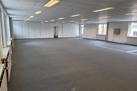 En stor, tomt kontorrum med hvide vægge, højt gulv og store vinduer. Der er et højt loft med mange lyspunkter og en dør i