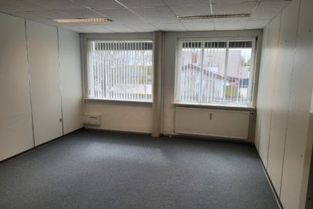 Et tomt kontor med to vinduer, hvor der er gardiner ned. Der er et radiator under hvert vindue. Der er to hvide vægge med