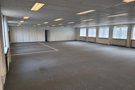 Tomt kontorrum med lyse hvide vægge, højt til loftet, lysekroner, gulvbelægning, vinduer med persienner og to rækker