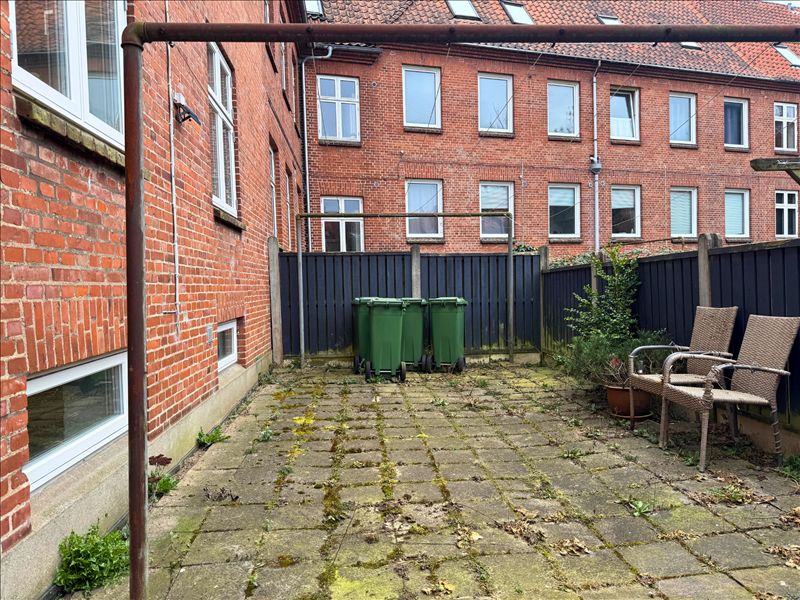 Smuk baghave med lyserøde mursten, to grønne skraldespande, en blomsterpotte og to sæder under et metalrørs stel