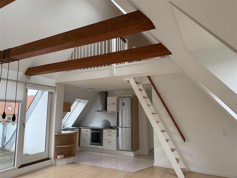 Loftsrum med lyse loftsbjælke og åbne ruder, gulvtæppe og lyse møbler i lyse farver