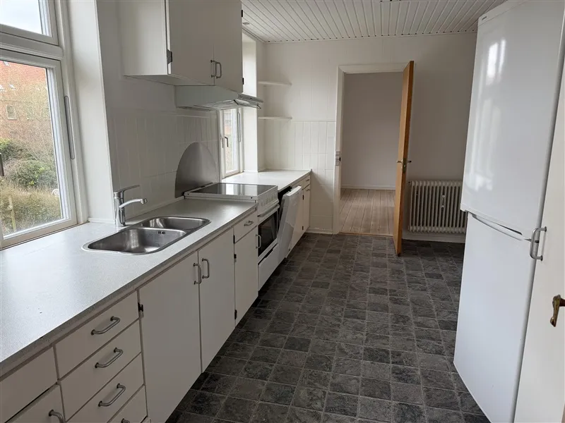 Et køkken med en grydefad, en ovn og en køleskab samt højloftede og hvide vægge