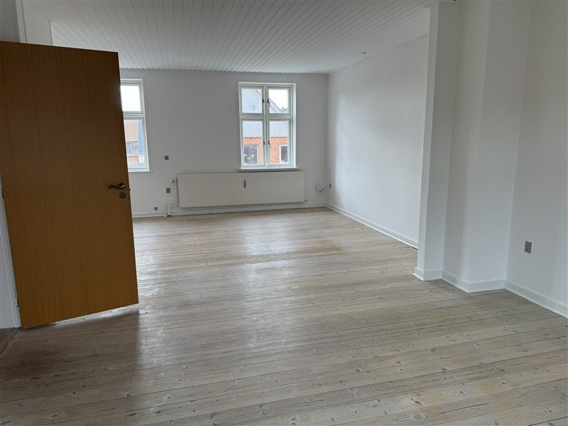 En tom stue med lysegul trægulv og hvide vægge og lofter, et åbent træpanel og to vinduer med hvide ruder