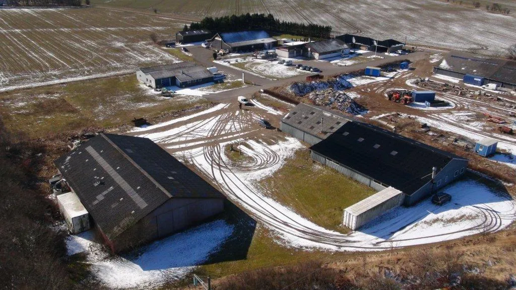 Vinterskov med sne og spor i sneen, bygninger og veje, og en gård med bilparkering og maskiner.