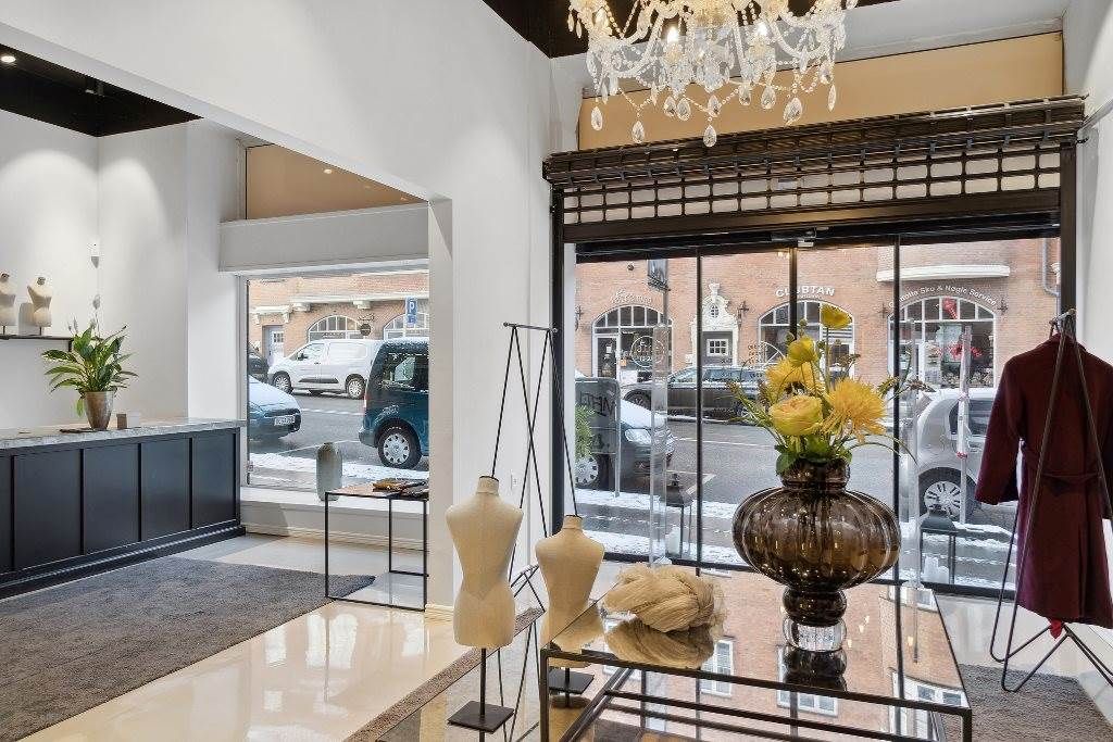 En indgangsparti til en butik med vinduer, der reflekterer biler og en bygning. En marmorbord med en vase med gule blomster