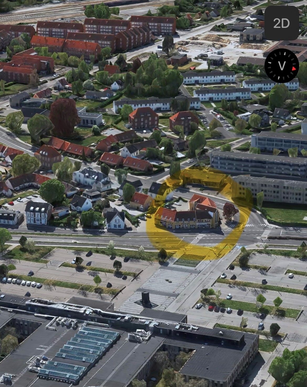 Flyfoto af bydel med røde tegltag, lyseblå himmel, og en stor gul cirkel markerer en bygning i midten af vejen