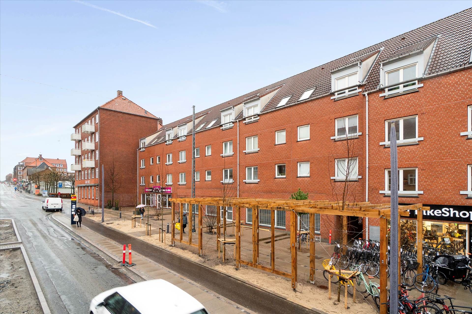 Bred vej med parkeringspladser og gågader med gadebelys og søjler samt hus med lejligheder og butikker med udsalgsvinduer og