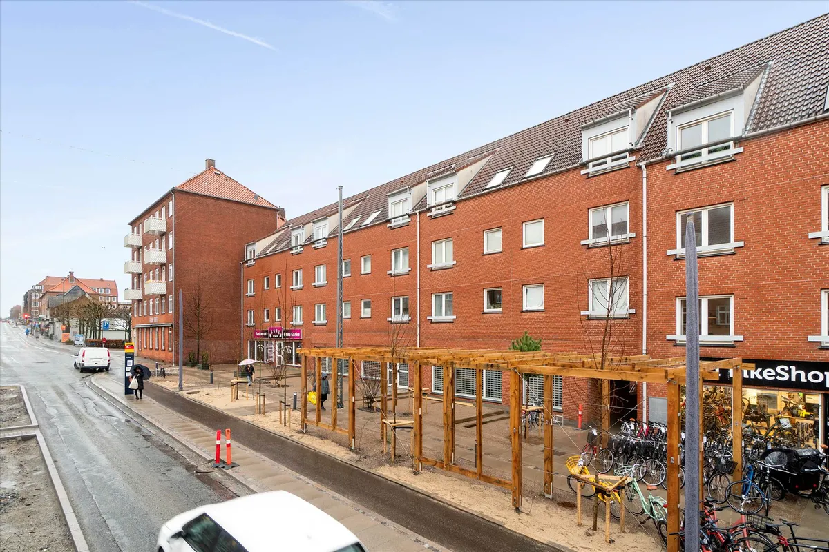 Bred vej med parkeringspladser og gågader med gadebelys og søjler samt hus med lejligheder og butikker med udsalgsvinduer og