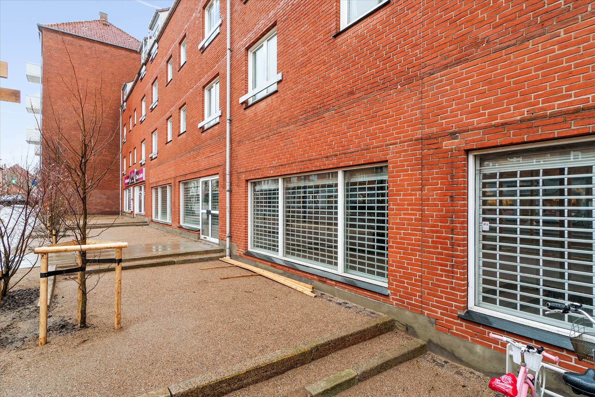 Rummeligt lejemål med store ruder og åbent område foran, hvor en lille træ er plantet, og en pink cykel er parkeret