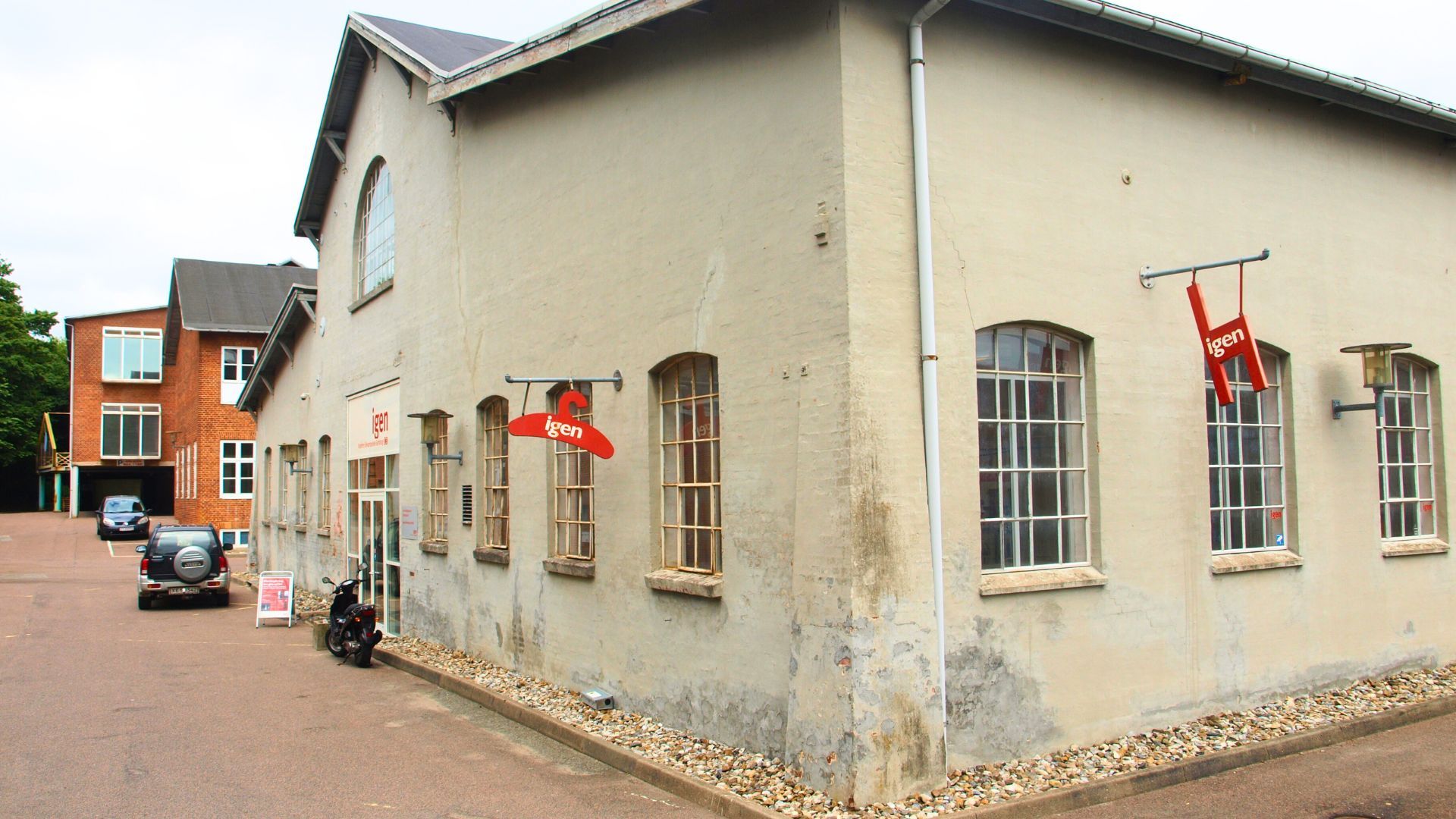 Butiksbygning med skilte og røde henvisningsskilte, parkeringsplads med bil og motorcykel, bøsser og grønne træer i