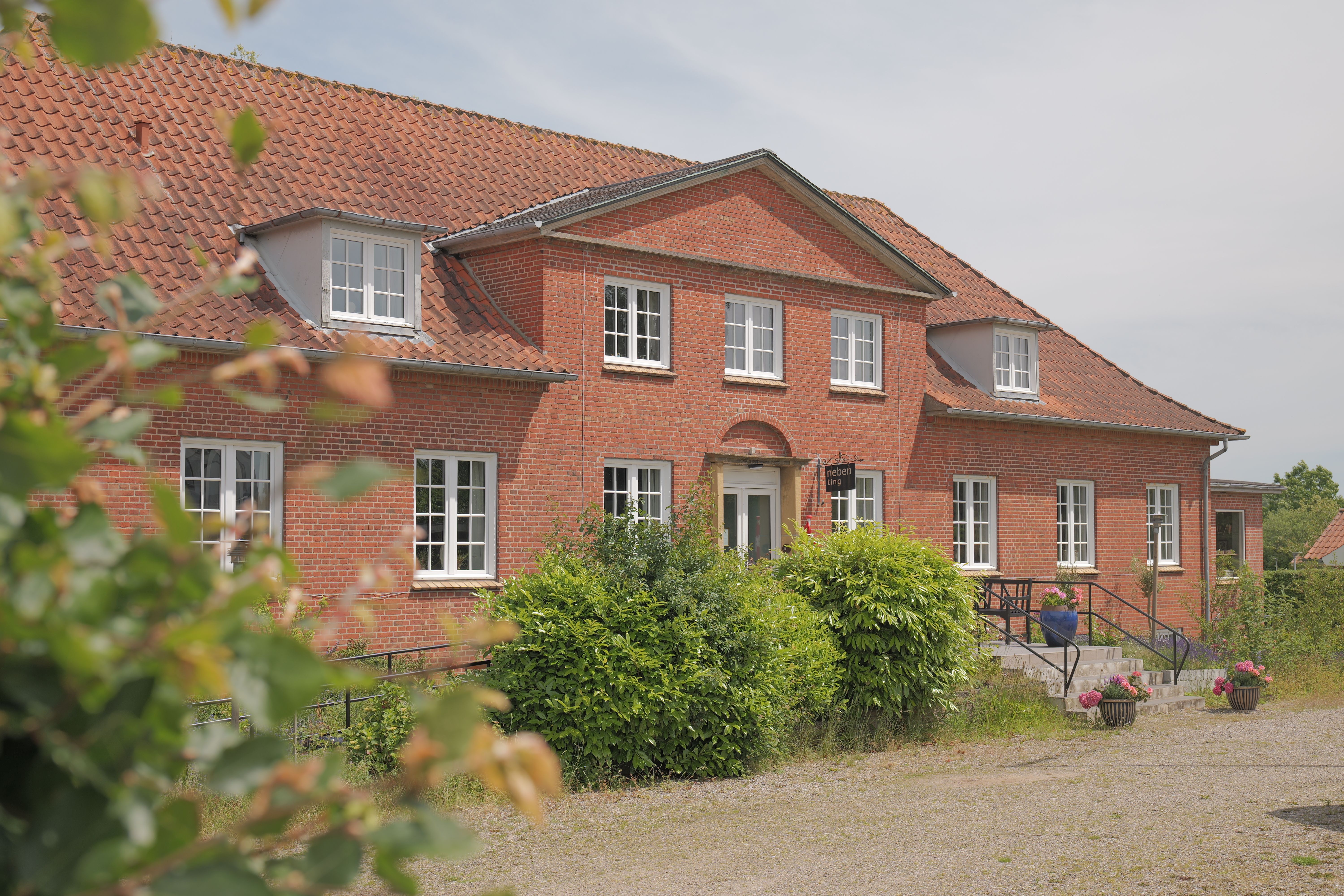 Rød teglstens bygning med mange vinduer og to blomsterpotter på trappen. Et schild med tekst "neben ting" hængende over