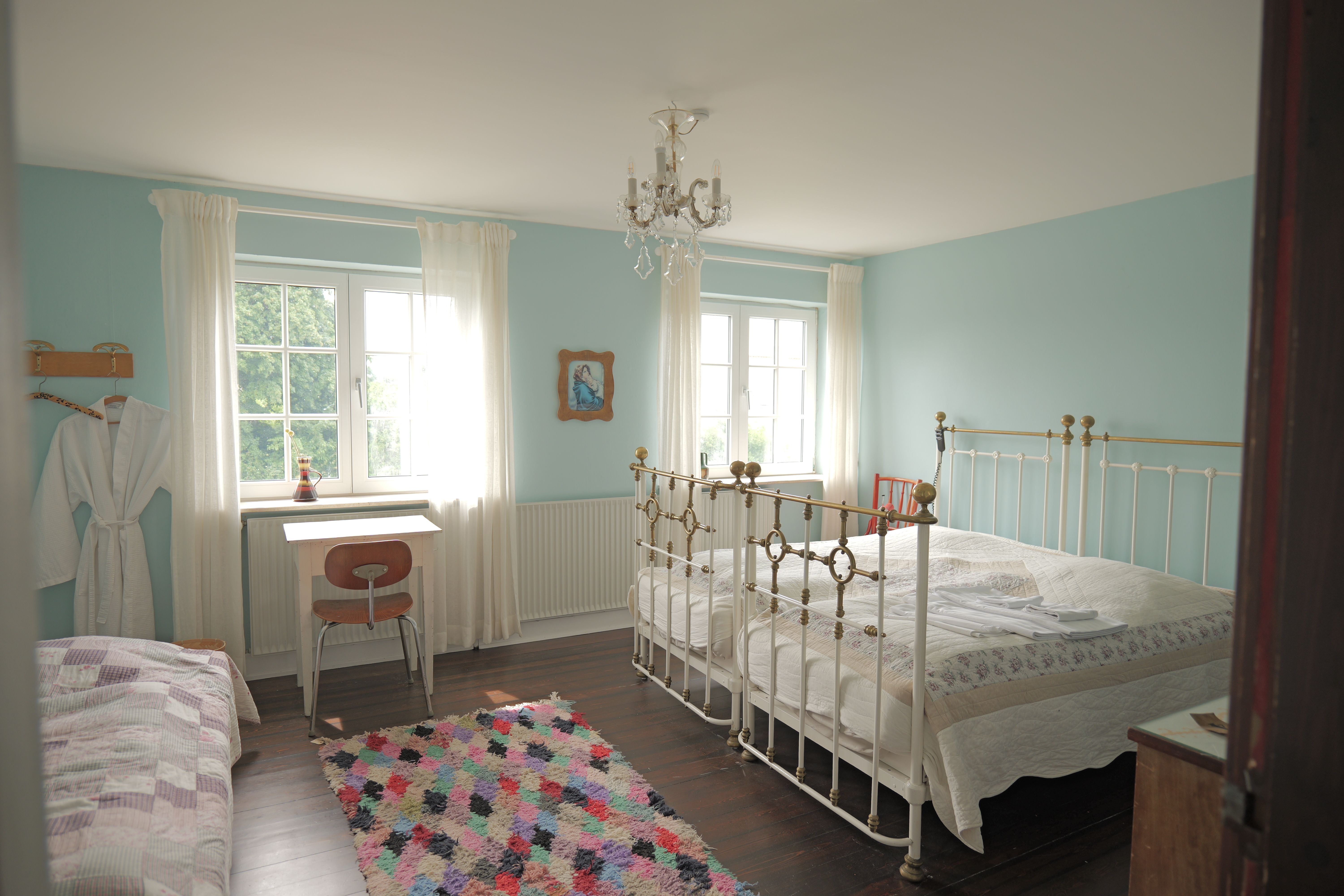 To-personers seng med høj stol og skrivebord i soveværelse med lyseblå vægge og lysegrå gardiner, hvor der er et billede