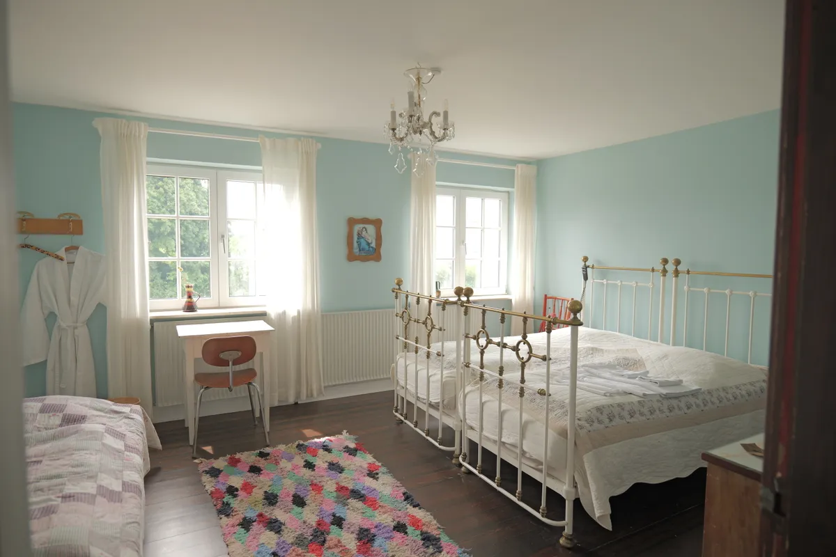 To-personers seng med høj stol og skrivebord i soveværelse med lyseblå vægge og lysegrå gardiner, hvor der er et billede