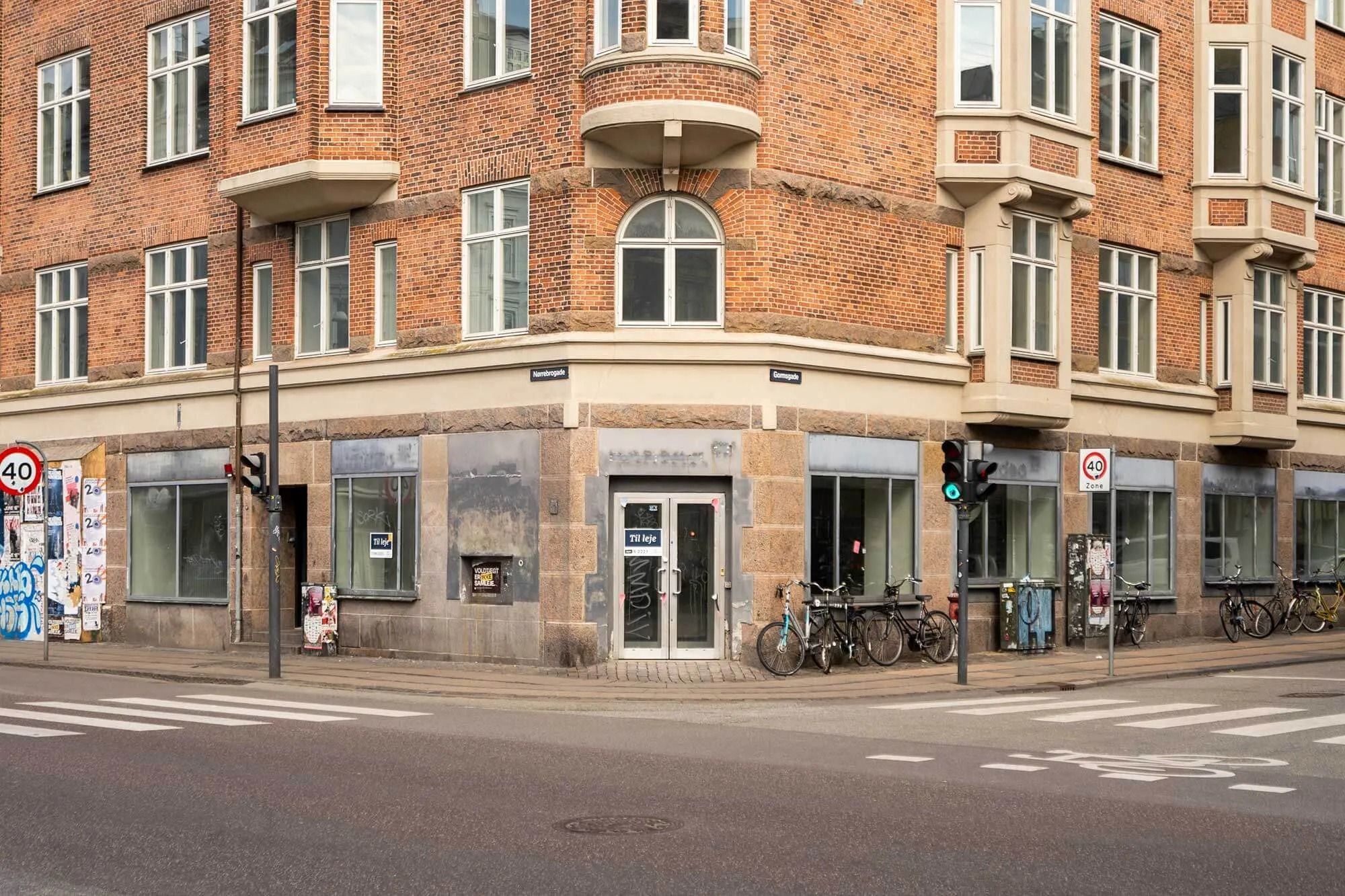 Rødstensbygning med mange vinduerne, en 40 km/t-skilt og cykler parkeret udenfor og et skilt med teksten "Til leje"