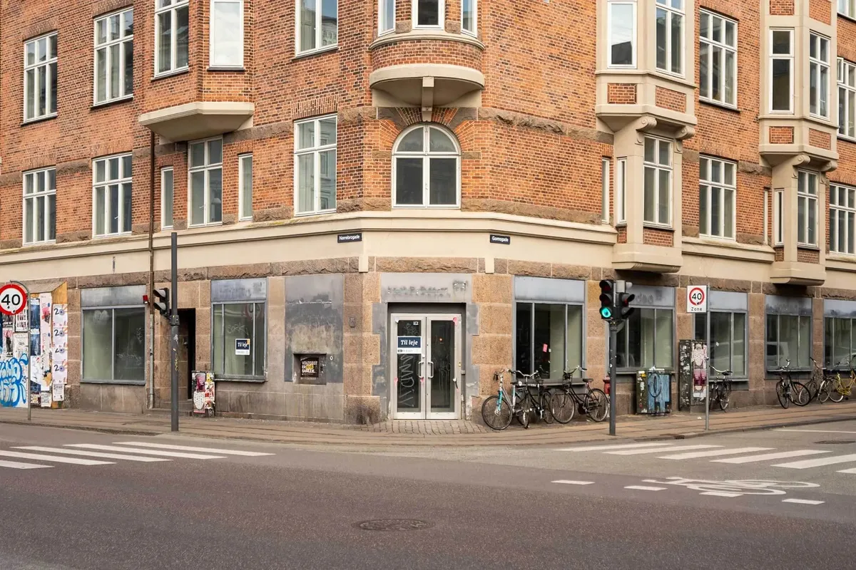 Rødstensbygning med mange vinduerne, en 40 km/t-skilt og cykler parkeret udenfor og et skilt med teksten "Til leje"