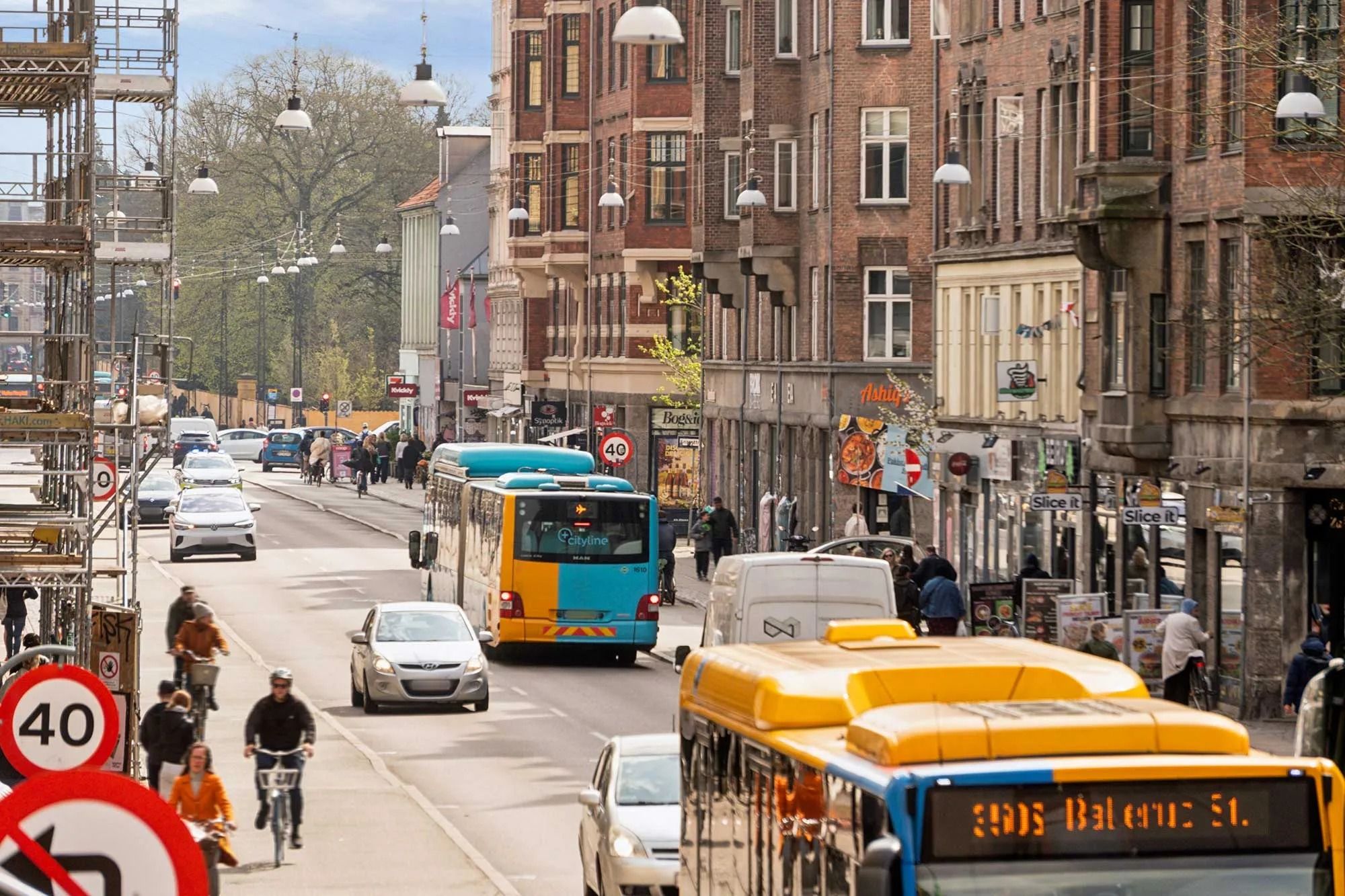 En travl vej med biler og busser. Forskellige reklamer og skilte. Mennesker og cyklister på fortovet. Bygninger med røde