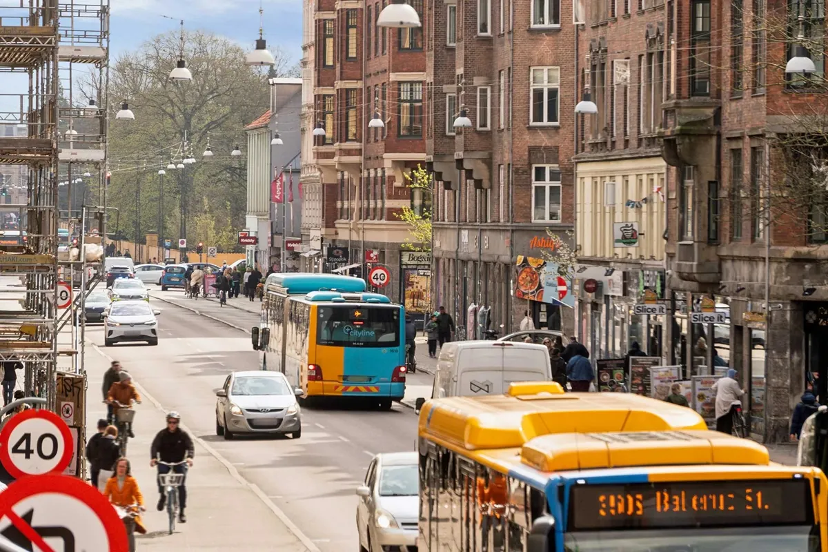 En travl vej med biler og busser. Forskellige reklamer og skilte. Mennesker og cyklister på fortovet. Bygninger med røde