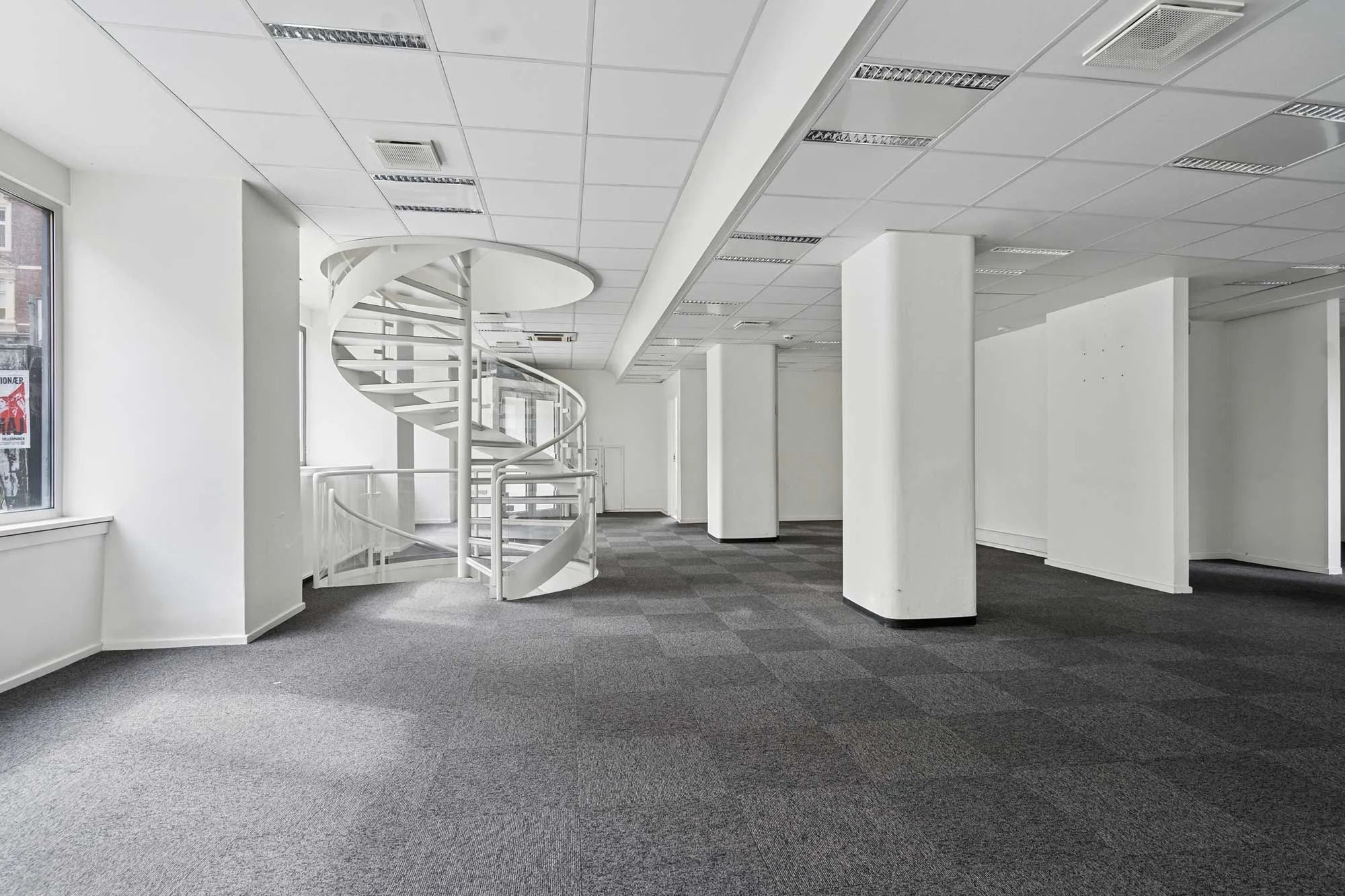 Indenfor en tom kontoret er der en hvid spiraltrappe og fire hvide søjler foran et vindue med et reklameplakat