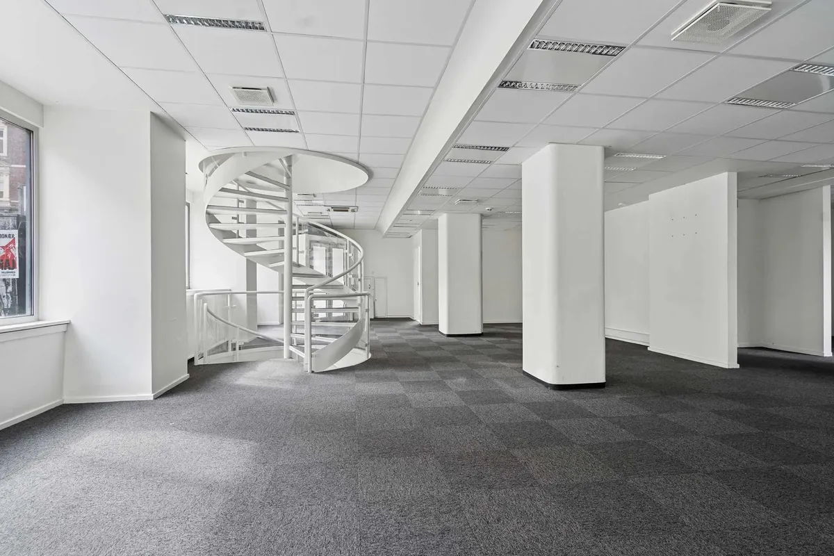 Indenfor en tom kontoret er der en hvid spiraltrappe og fire hvide søjler foran et vindue med et reklameplakat