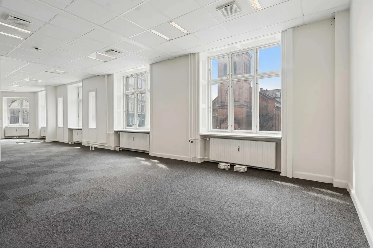 Et tomt kontor med lysegrå gulvbelægning og hvide vægge samt vinduer og radiatorer med udsigt til en bygning udenfor