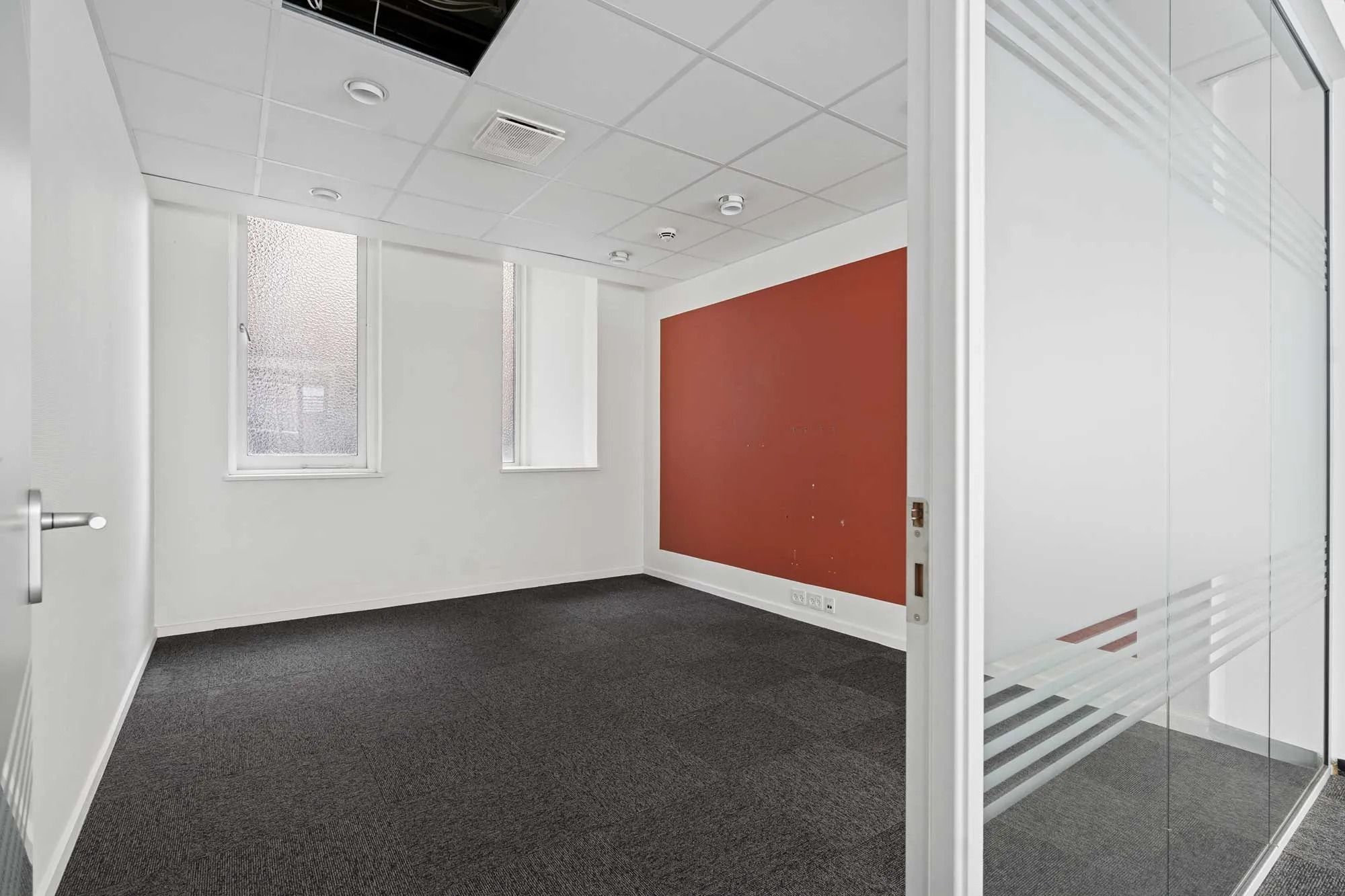 En tom, velholdt kontorlokal med hvide vægge, rødt panel og gulvbelægning, to vinduer og en hvid glasdør.