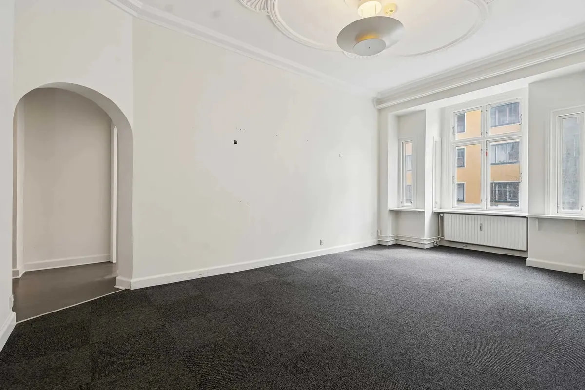 Et stort, tomt værelse med hvide vægge og lysegule gulvbelægning, to store vinduer med hvide ruder og en hvid runde