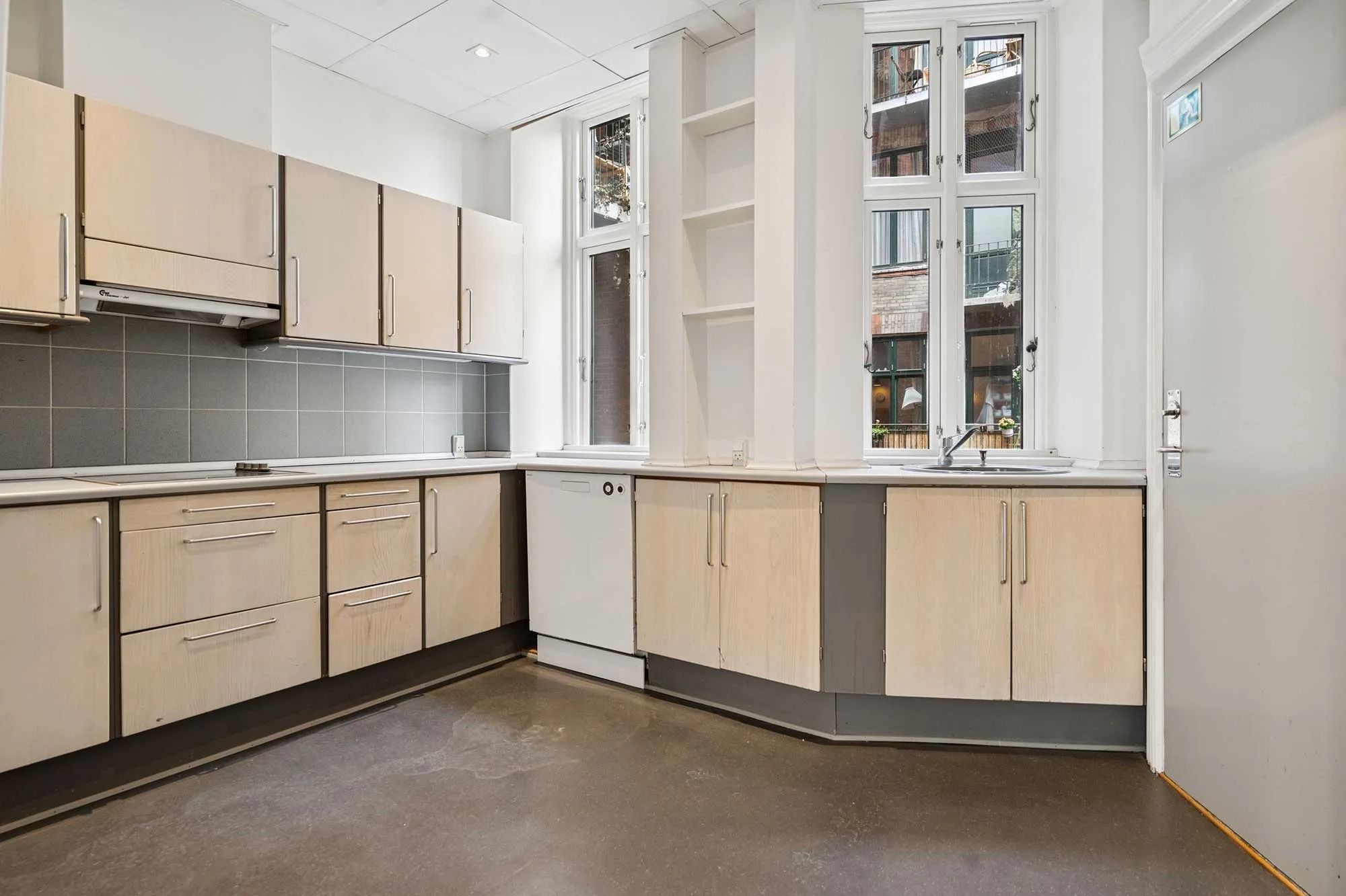 Et moderne køkken med høje hvide vægge, lyse træskabe, rustfrit stål sink, og store vinduer med udsigt til byen.